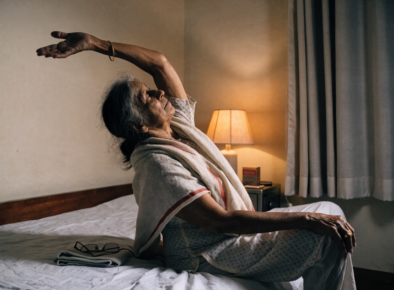 An older Indian woman doing a calming evening stretch on her bed, in a simply furnished bedroom with warm lamp light